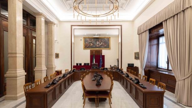 Formal council chamber with U-shaped wooden desks, flags at the front, a round chandelier, marble columns, and heavy curtains.