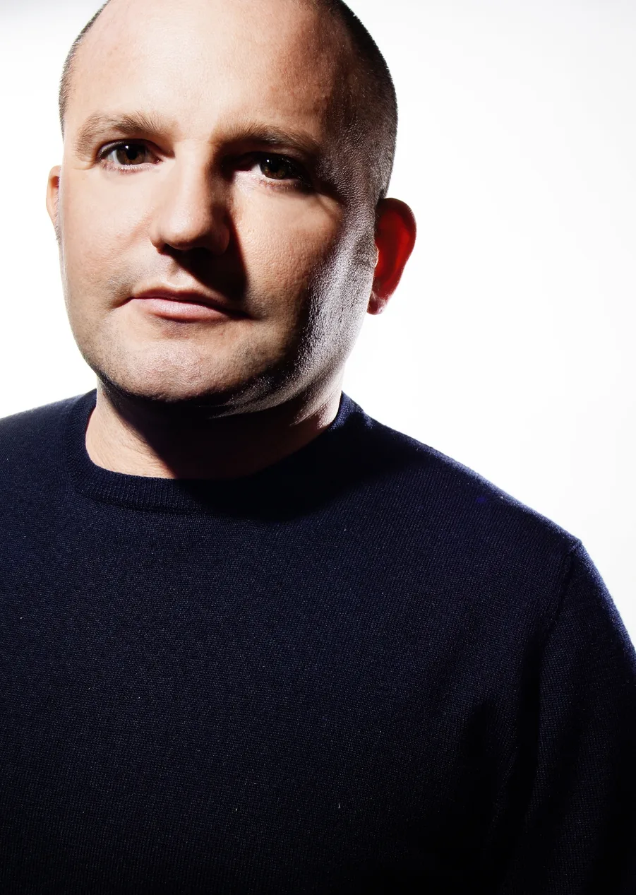 Close-up portrait of a man with a shaved head wearing a dark sweater against a white background.